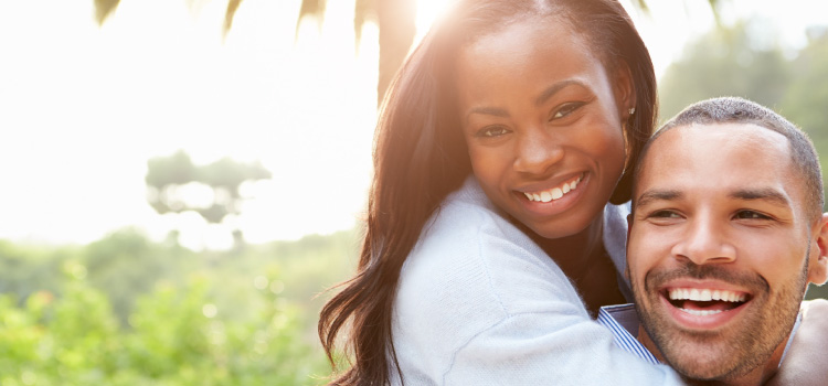 woman smiling and riding piggy back on man smiling, outside with greenery in background