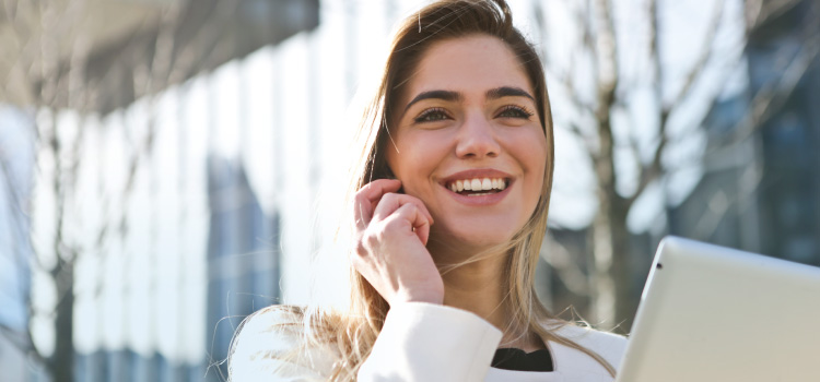 blonde woman with white teeth, holding hand up next to face, smiling, standing outside building