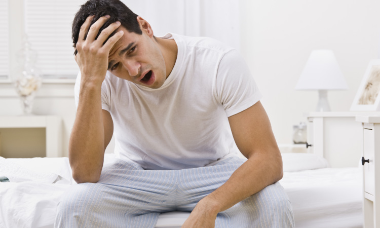 Brunette man with obstructive sleep apnea sits on the edge of his bed yawning in his pajamas