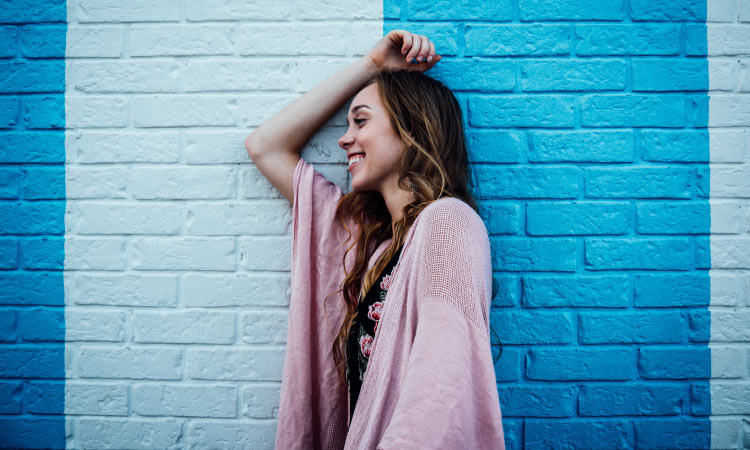 Brunette woman wearing a pink cardigan smiles as she leans sideways against a brick wall painted 2 colors of blue