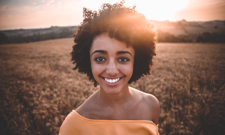 Brunette young woman who uses floss every day smiles while wearing a yellow blouse and standing in a wheat field