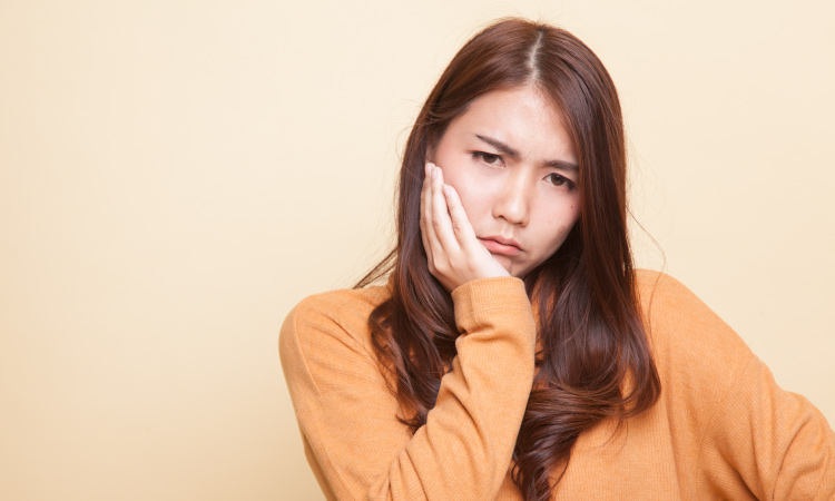 Brunette woman wearing an orange sweater cringes and touches her cheek due to sensitive teeth