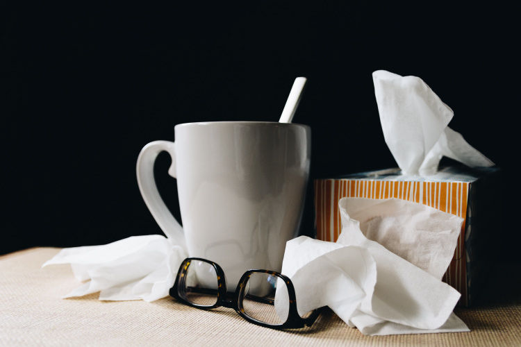 A box of tissues next to a white mug, glasses, and used tissues by someone with a cold