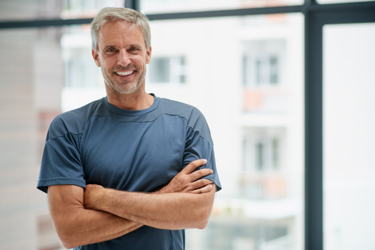 Middle-aged man smiles with dental implants while wearing a gray blue shirt and folding his arms