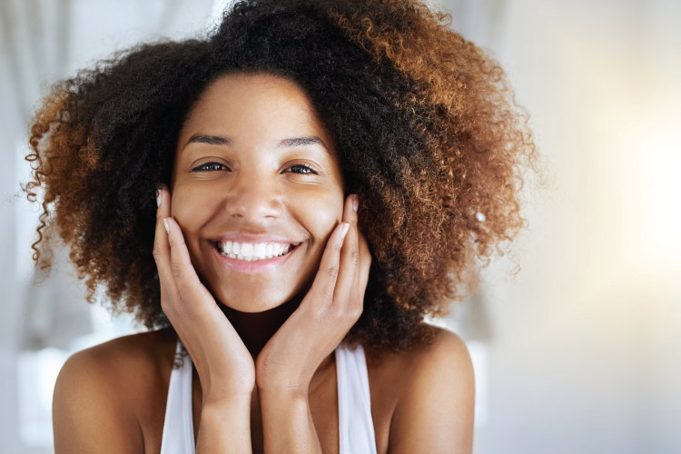 Curly brunette woman with dental veneers smiles brightly while touching her cheeks and wearing a white tanktop