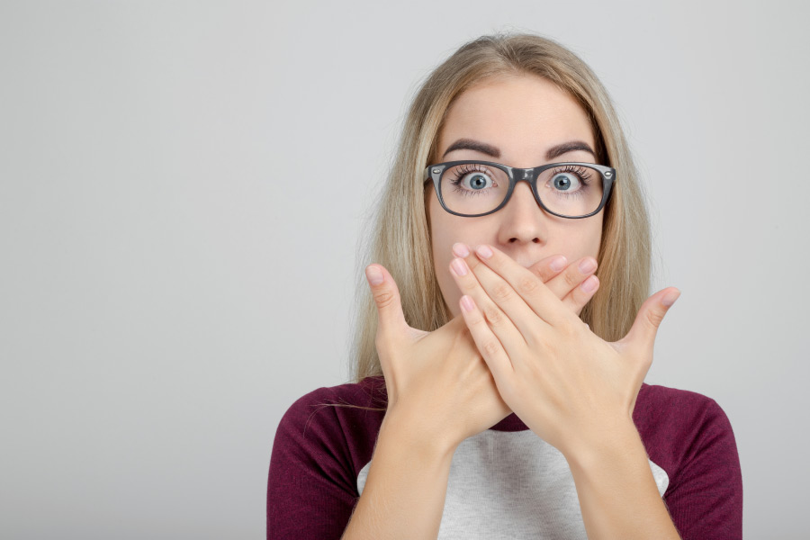 young woman wearing glasses covers her mouth with both hands to hide her sore gums