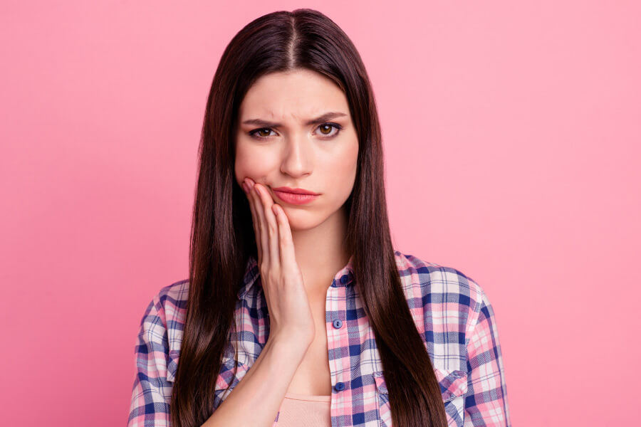 Brunette woman cringes in pain and touches her cheek due to sensitive teeth against a pink wall