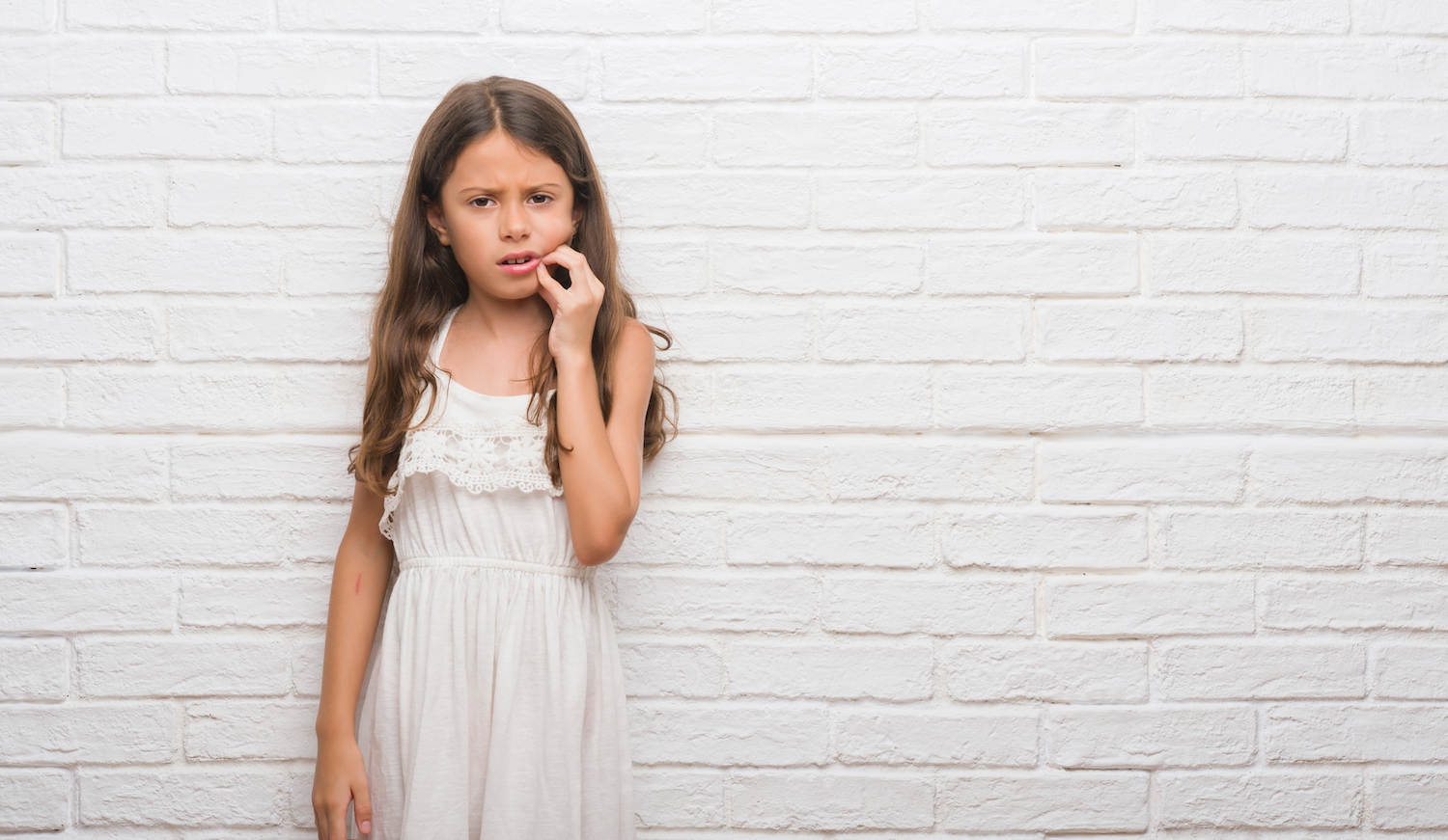 Brunette little girl in a white jumper cringing in pain and touching her cheek due to a toothache