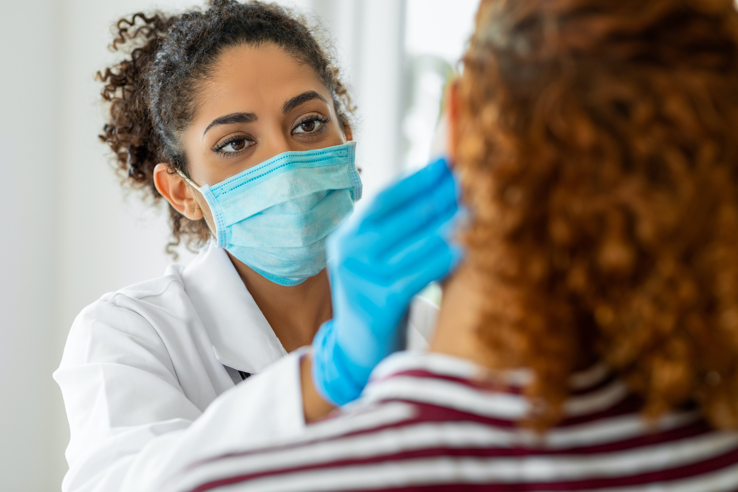 Closeup of a dentist wearing blue gloves and a blue mask as she palpates a patient's neck for lumps during an oral cancer screening