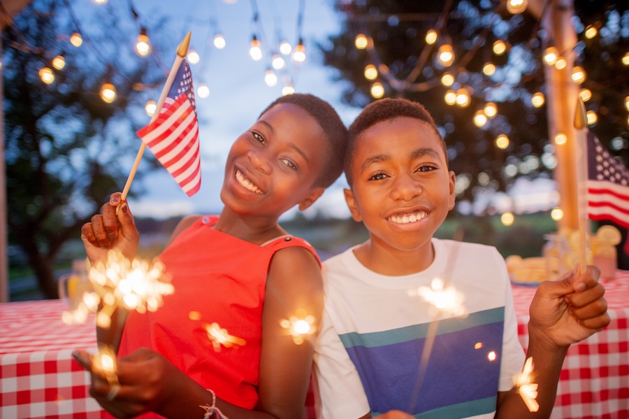two young kids celebrating july 4 with smiles, flags, and sparkers at a party, dental safety