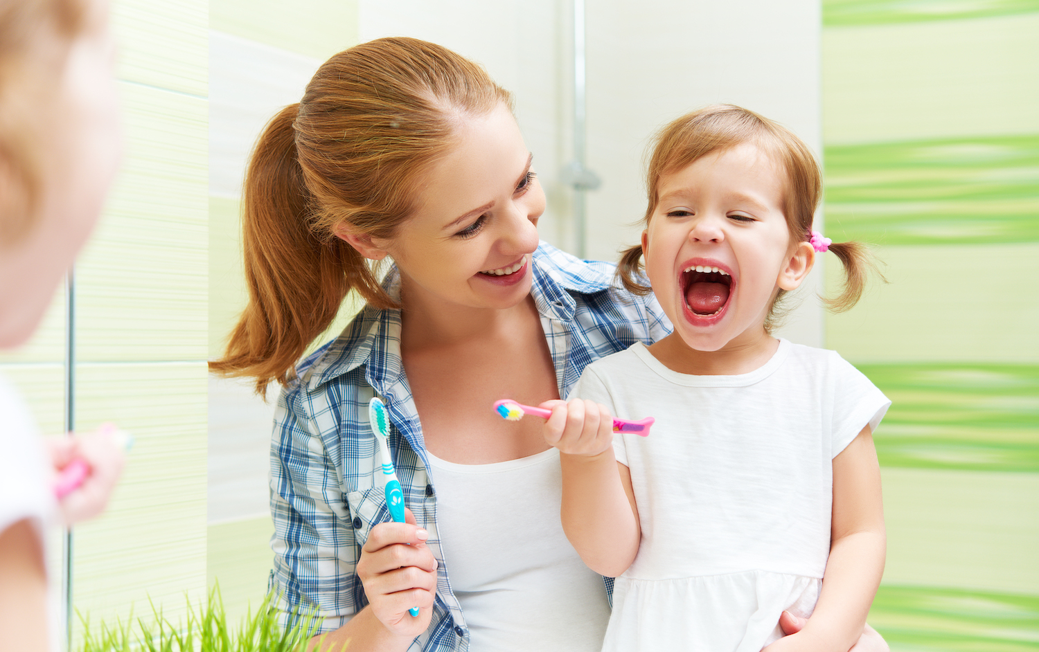 Woman smiles as she looks at her young daughter brushing her teeth in the bathroom