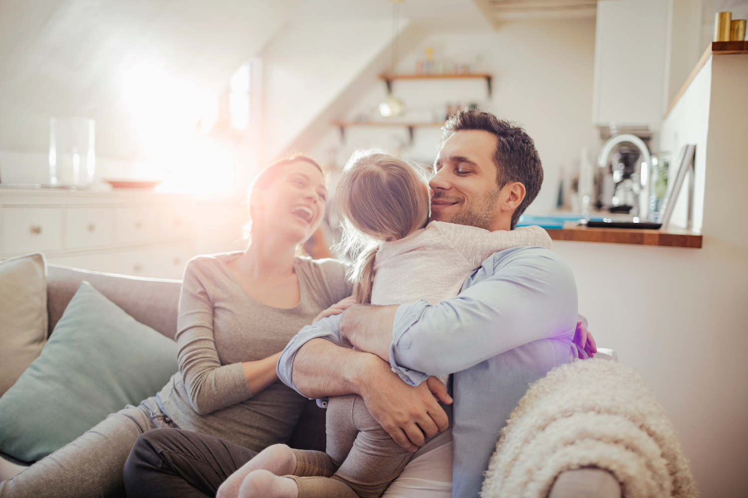Mom and Dad smile as they embrace their young daughter on the couch in their family room