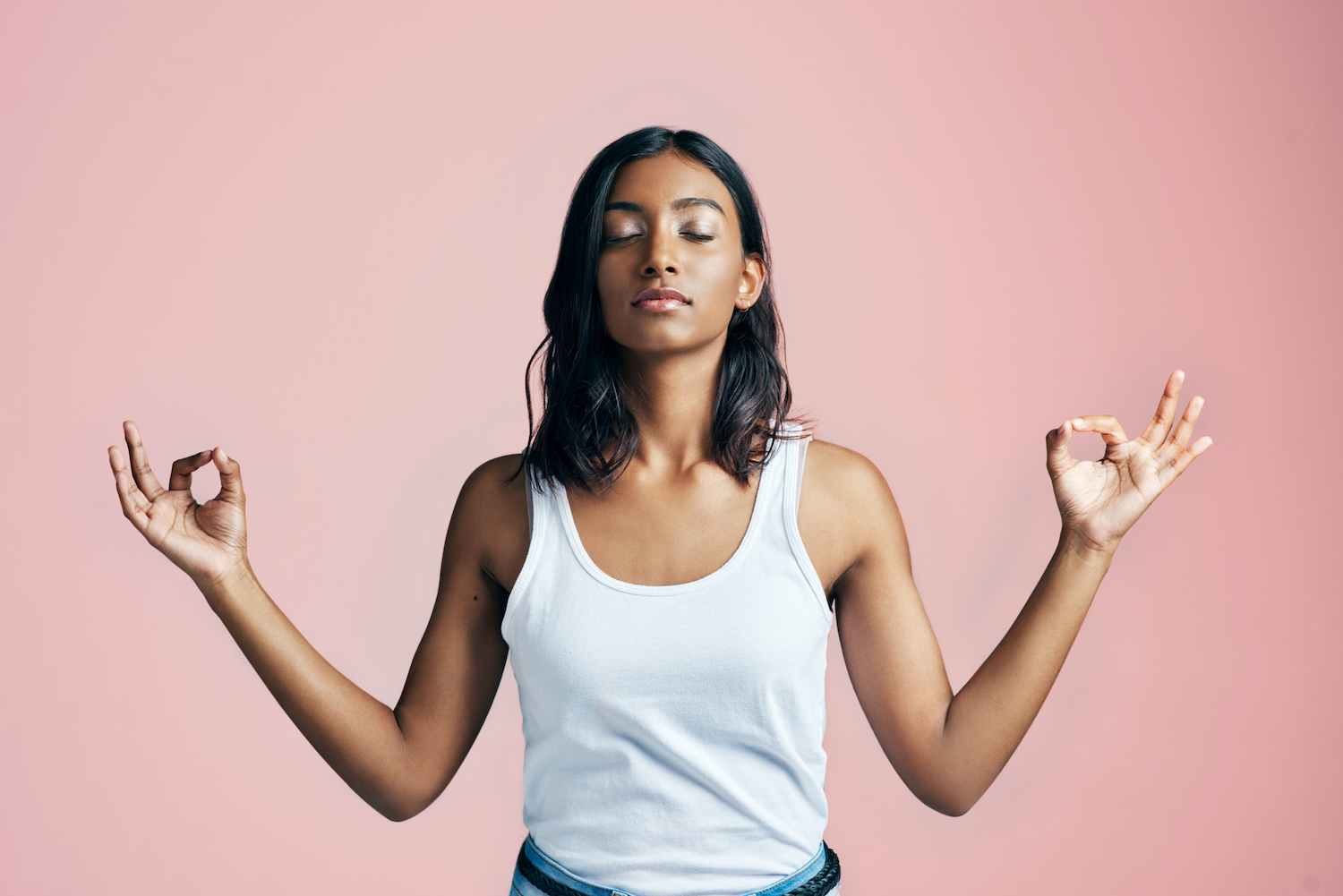 Brown woman in a white tanktop closes her eyes and holds her hands in a zen pose as she relaxes against a pink background