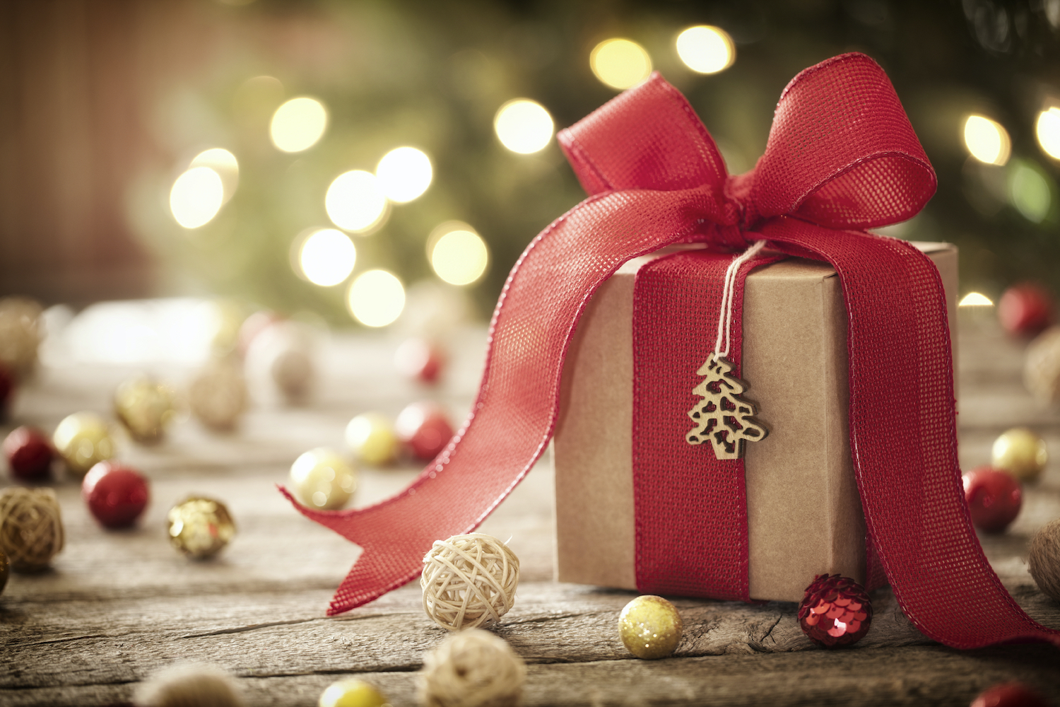 Closeup of a small holiday gift wrapped in brown paper and a red ribbon surrounded by festive balls by a Christmas tree