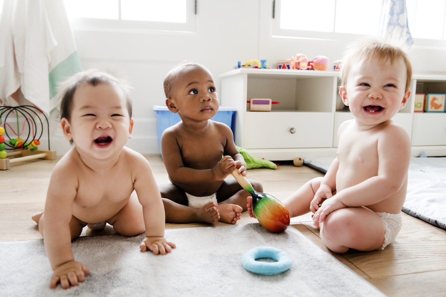 3 teething babies in their diapers smile as they play together on the floor with toys