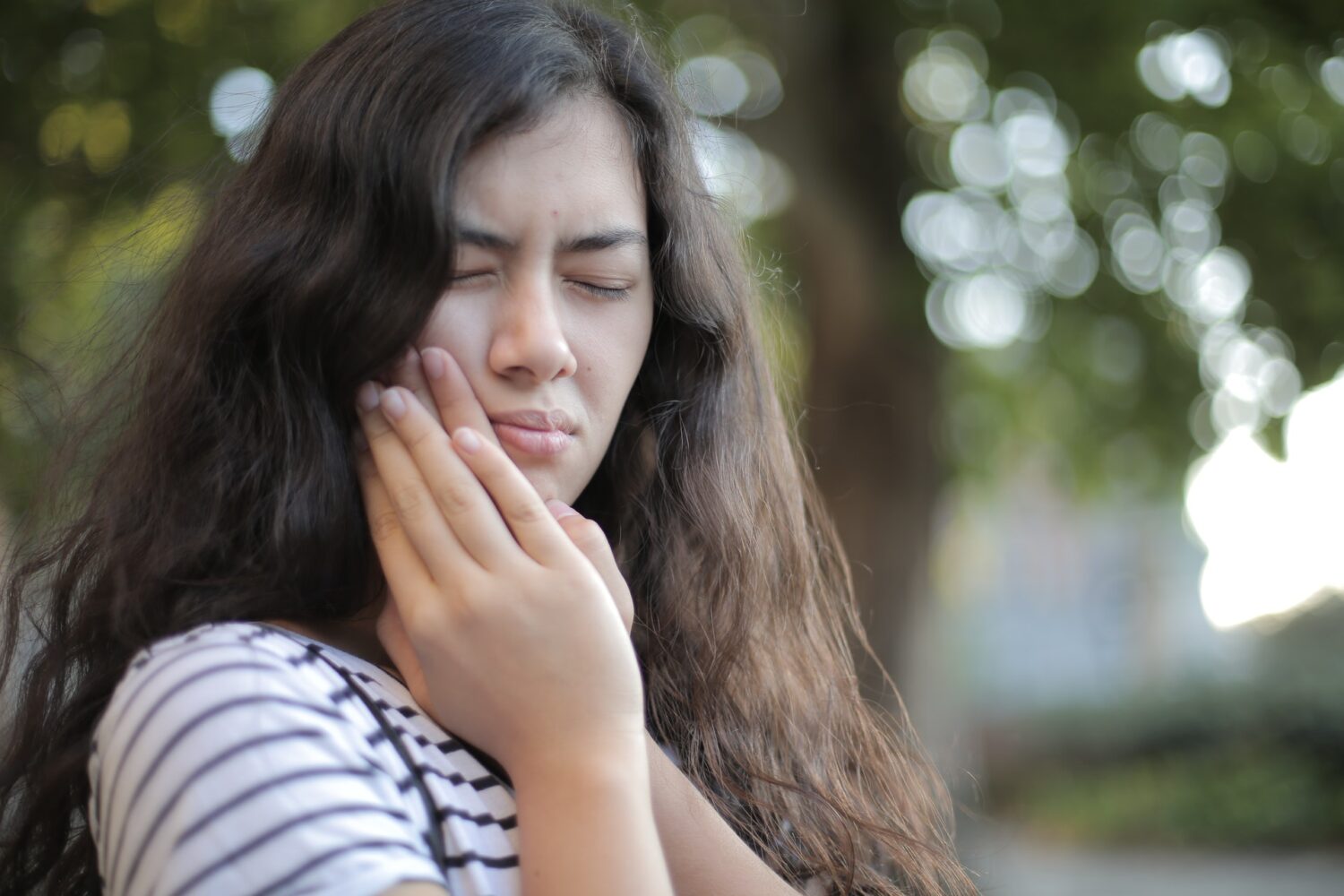 Brunette woman cringes in pain and holds her cheek due to tooth sensitivity