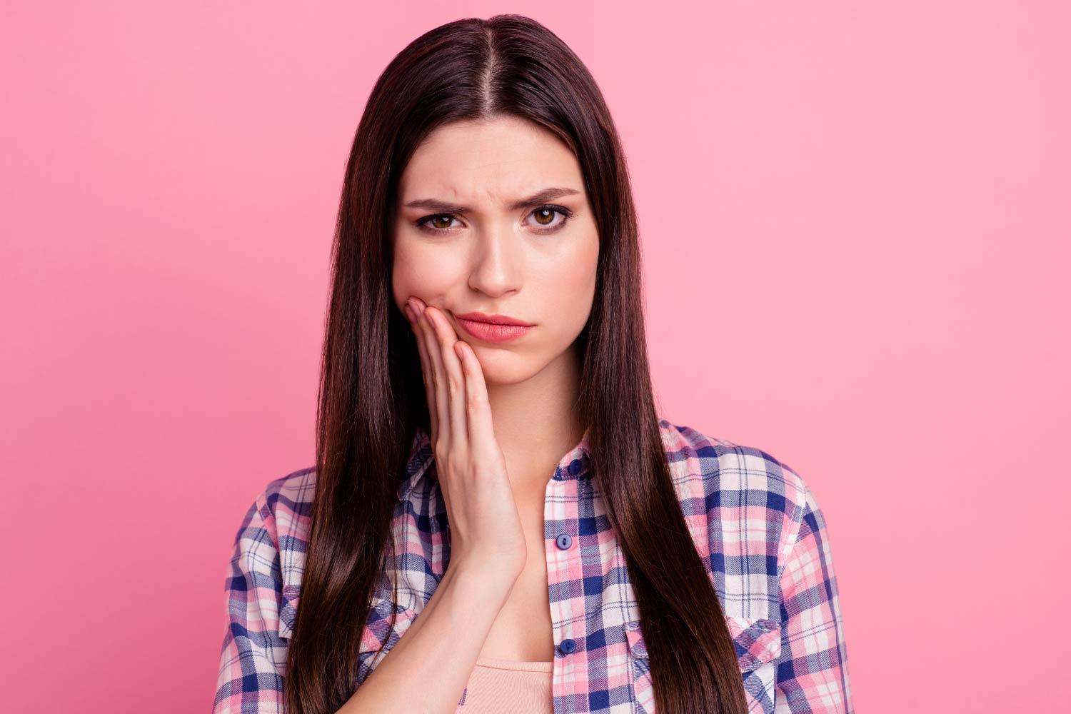 Brunette white woman in a plaid t-shirt against a pink wall cringes in pain and touches her cheek due to a toothache